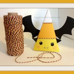 May include: A yellow and orange paper cone shaped like a candy corn with a white top and black bat wings. The cone has a cute cartoon face with black eyes, a red mouth, and two small fangs. A spool of orange and brown twine is in the background.