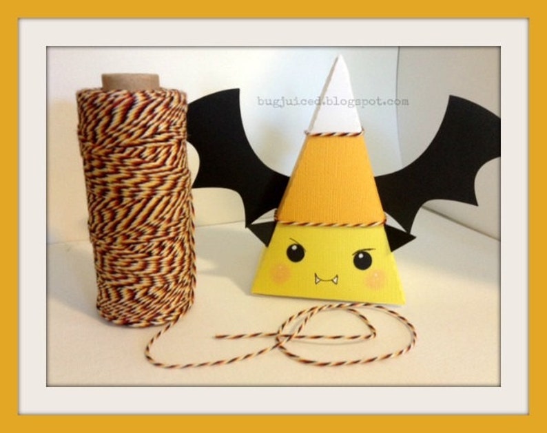 May include: A yellow and orange paper cone shaped like a candy corn with a white top and black bat wings. The cone has a cute cartoon face with black eyes, a red mouth, and two small fangs. A spool of orange and brown twine is in the background.