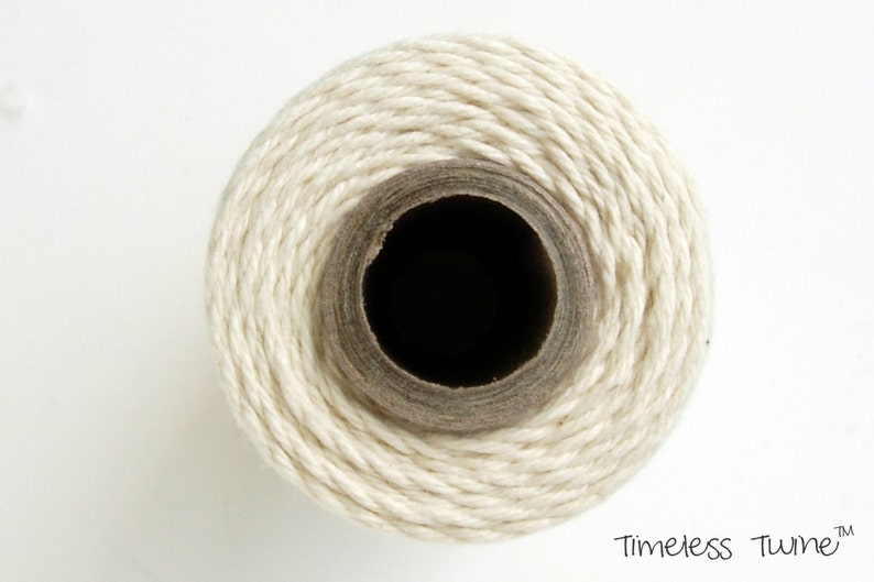 May include: A close-up of a spool of natural white twine. The twine is wound tightly around a cardboard core.