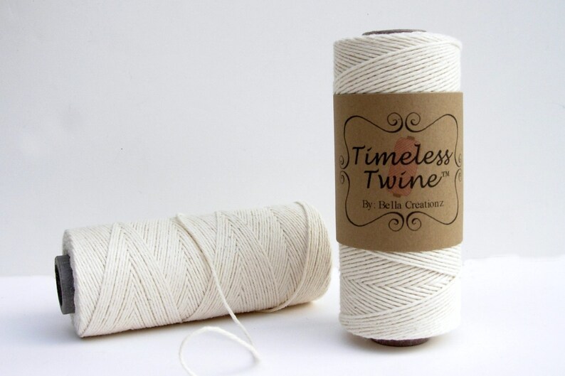 May include: Two spools of natural white cotton twine. The twine is wound around cardboard spools with brown paper labels. The label on the spool in the foreground reads "Timeless Twine&trade; By: Bella Creations."