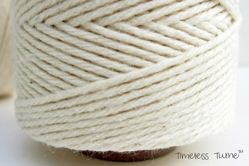 May include: A close-up of a spool of natural white cotton twine. The twine is tightly wound and has a smooth, even texture. The spool is labeled "Timeless Twine".