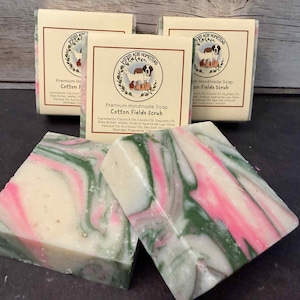 May include: Four bars of handmade soap with a swirling pattern of pink and green on a white base. The soap is labeled "Cotton Fields Scrub" and features a logo with a dog and a sheep. The soap bars are on a dark surface.