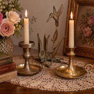 Puede incluir: Dos candelabros de latón con velas cónicas encendidas. Uno tiene una base ancha y un asa, mientras que el otro presenta un diseño más sencillo. Entre ellos se encuentra una escultura de metal color bronce de un colibrí sobre un tallo.