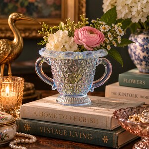 May include: A light blue iridescent glass vase with a diamond-patterned body and two curved handles, featuring a scalloped rim. The vase is filled with a bouquet of pink ranunculus, white hydrangea, and small white daisies, resting on books.