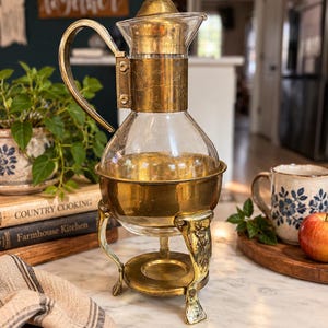 May include: A clear glass carafe with a brass lid and handle, featuring etched floral details. The carafe rests in a matching brass stand with three curved legs and a circular base, designed to hold the glass vessel securely in an upright position.