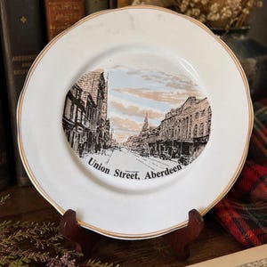 Puede incluir: Un plato decorativo de cerámica blanca con borde dorado que presenta una ilustración en blanco y negro de una escena callejera. El texto Union Street, Aberdeen está impreso en negro en la parte inferior de la imagen central, que muestra edificios.