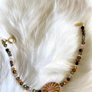 May include: A delicate bracelet with a pink and gold sunburst charm, featuring a mix of brown, black, and clear glass beads.