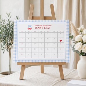 May include: A baby shower calendar on a wooden easel. The calendar has a blue gingham border and the text "GUESS THE ARRIVAL OF BABY LEO". A red heart marks the 14th. White flowers and greenery are in the background.