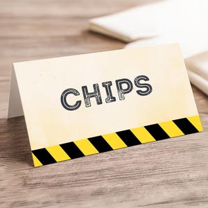 May include: A cream-colored card with the word "CHIPS" in black, outlined letters. The bottom of the card features a black and yellow hazard stripe design. The card is propped up on a wooden surface.