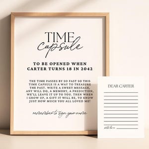 May include: A framed print with the words "TIME capsule" in elegant script. The text below reads "TO BE OPENED WHEN CARTER TURNS 18 IN 2042." A smaller card with lines and the text "DEAR CARTER" is next to the frame. The frame is light brown.