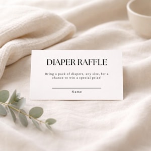 May include: A white card with the text "DIAPER RAFFLE" in a bold, elegant font. Below, the card reads, "Bring a pack of diapers, any size, for a chance to win a special prize!" with a line for the name. The card is on a cream-colored fabric.