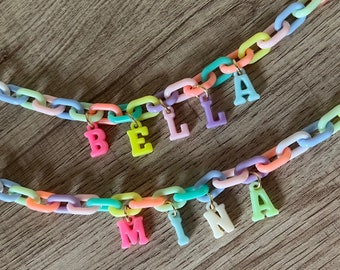 May include: Two colorful plastic chain necklaces with letter charms spelling out the names "BELLA" and "MINA".