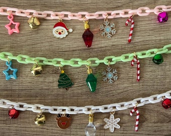 May include: Three Christmas-themed necklaces with different colored chains. The top necklace has a pink chain with charms including a star, bell, Santa Claus, light bulb, snowflake, and candy cane. The middle necklace has a green chain with charms including a star, bell, Christmas tree, light bulb, snowflake, and candy cane. The bottom necklace has a white chain with charms including a bell, bell, reindeer, light bulb, snowflake, and candy cane.