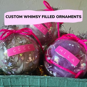 Puede incluir: Tres adornos de vidrio transparente llenos de diferentes materiales. El adorno de la izquierda está lleno de purpurina blanca y plateada, pequeñas flores blancas y una etiqueta rosa con el nombre "Cece". El adorno del medio está lleno de hilo rosa y blanco y una etiqueta rosa con el nombre "Mila". El adorno de la derecha está lleno de hilo rosa y blanco y una etiqueta rosa con el nombre "Lily". Los adornos están en una cesta verde.