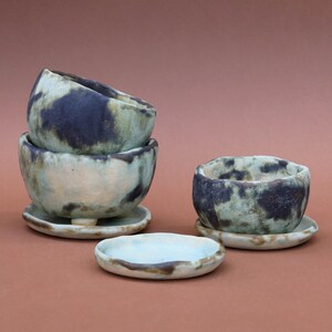 May include: Handmade ceramic bowls and saucers in shades of blue, green, and white. The bowls have a speckled design and are stacked in a group of three, with a single bowl and saucer set nearby. The small saucers are round and have a raised edge.