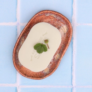 Pode incluir: Uma saboneteira de cerâmica ovalada feita à mão com um esmalte cor de cobre. Uma barra de sabão de cor clara repousa no interior, adornada com uma pequena folha verde e um pequeno botão. A saboneteira está sobre uma superfície de azulejos azuis.