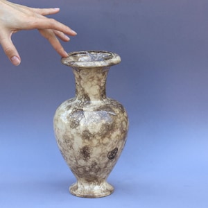 Könnte beinhalten: Eine dekorative Vase mit einem gesprenkelten Muster in Beige- und Brauntönen. Die Vase hat einen breiten Sockel, einen schlanken Hals und einen ausgestellten Rand. Die Gesamthöhe beträgt ca. 25 cm.