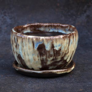 May include: A small, round ceramic planter with a matching saucer. The pot features a reactive glaze with a blend of dark brown, cream, and light blue colors that drip down the exterior. The rim is uneven and textured, showing a dark brown base.
