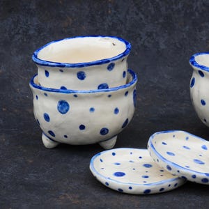 Op de afbeelding: Handgemaakte keramische kommen en schotels. Het witte aardewerk heeft blauwe stippen en een blauwe rand. Eén kom is op een andere gestapeld en er zijn verschillende schotels zichtbaar. De kommen hebben kleine voetjes.