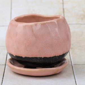 Op de afbeelding: Een keramische plantenpot met een afgeronde vorm en een bijpassende schotel. De pot is voornamelijk roze met een zwarte band bij de basis. De schotel is ook roze. Dit decoratieve item is geschikt voor kleine planten.