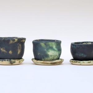 May include: Three small, handmade ceramic pots with matching saucers. The pots are dark blue with green and yellow accents, and the saucers are a light beige color. The pots are of varying sizes, perfect for small plants or succulents.