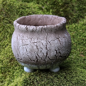 Peut inclure: Un petit pot de fleurs en céramique ronde avec une finition extérieure texturée et craquelée dans des tons gris et marron. Le récipient présente une large ouverture sur le dessus et repose sur trois petits pieds arrondis, offrant un aspect rustique.