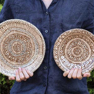Peut inclure: Deux assiettes en céramique faites à la main avec des motifs bruns complexes. Les assiettes présentent des cercles concentriques et des motifs de points, créant un aspect artistique unique. Les assiettes sont tenues contre une chemise bleu foncé et un feuillage vert.
