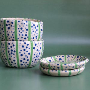 May include: Two stacked white ceramic bowls and two small plates. The bowls and plates are decorated with blue polka dots and green vertical stripes. The background is a solid, muted green.