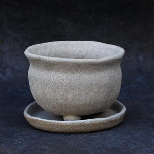 May include: A light gray, speckled ceramic planter with a rounded, bulbous body and a flared rim. The planter features a textured surface and rests on small, integrated feet, accompanied by a matching circular saucer with a slightly raised edge.