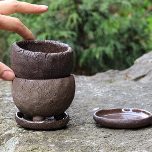 Puede incluir: Juego de tazas de té de cerámica marrón oscuro hechas a mano. El juego incluye una taza de té pequeña y redonda, una base a juego y un platillo pequeño. La taza de té tiene una superficie texturizada y un aspecto rústico.