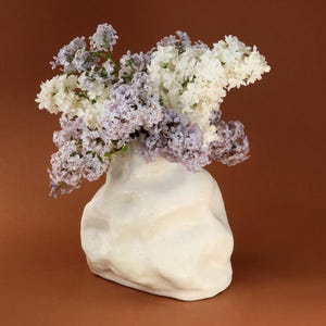 May include: A white ceramic vase with an organic, textured shape, filled with a bouquet of purple and white lilac flowers.