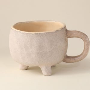 Puede incluir: Una taza pequeña de cerámica blanca con una superficie texturizada y tres patas. La taza tiene una forma redondeada y un asa.