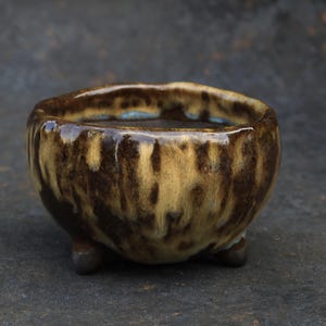 May include: A small, round ceramic bowl featuring a glossy glaze with a mottled pattern of dark brown and cream colors. The bowl has an uneven, organic rim and is supported by three short, rounded feet at the base, creating a tripod structure.