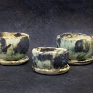 May include: Three ceramic bowls with matching saucers. The bowls have a rounded shape and are glazed in shades of green and black. The saucers are a lighter shade of green. The bowls are of varying sizes.