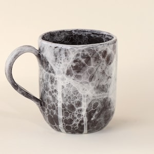 Könnte beinhalten: Eine Keramiktasse mit einem einzigartigen, strukturierten Design. Die Tasse hat einen dunkelgrauen Boden mit einem weißen, blasenartigen Muster und einem passenden Henkel. Der Innenraum ist weiß, und die Tasse hat eine zylindrische Form.