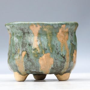 May include: A small, round ceramic planter with three short, light-colored feet. The exterior features a reactive glaze with mottled shades of forest green, dark teal, and patches of exposed tan clay, creating a textured, uneven appearance.