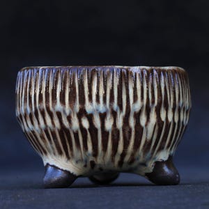 May include: A small, round ceramic planter with three short, dark brown feet. The exterior features a vertical ribbed texture with a two-tone glaze, alternating between deep dark brown and a creamy off-white colour that drips down the sides.