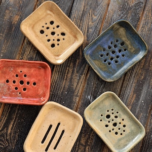 Puede incluir: Colección de jaboneras de cerámica hechas a mano en varios colores, incluyendo beige, rojo, azul y verde. Cada jabonera tiene un patrón único de agujeros o ranuras para el drenaje. Las jaboneras están dispuestas sobre una superficie de madera oscura.