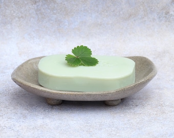 May include: A light green bar of soap with a small green leaf on top, resting in a speckled, oval-shaped soap dish. The dish has three small feet. The background is a light, textured surface.