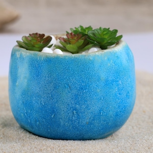 Op de afbeelding: Een kleine, ronde, turquoise keramische plantenbak gevuld met kleine groene en roodbruine vetplanten en witte kiezelstenen. De plantenbak heeft een getextureerd oppervlak en staat op een bed van zand, wat een natuurlijke, decoratieve presentatie creëert.