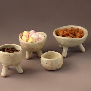 Pode incluir: Um conjunto de pequenas tigelas de cerâmica esbranquiçada com três pernas, cada uma preenchida com itens diferentes. Uma tigela contém grãos de café, outra marshmallows e uma terceira passas. A quarta tigela está vazia. Em fundo neutro.