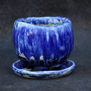 May include: A small, round ceramic planter with a vibrant cobalt blue glaze featuring white drip patterns. The pot has a textured, rippled surface, sits on three short feet, and includes a matching circular saucer base.