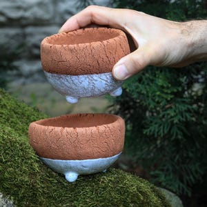 May include: Two small, handmade ceramic bowls. The top half of each bowl is a textured, reddish-brown color, while the bottom half is a light blue-gray. Each bowl has three small, white feet. One bowl is held by a hand.