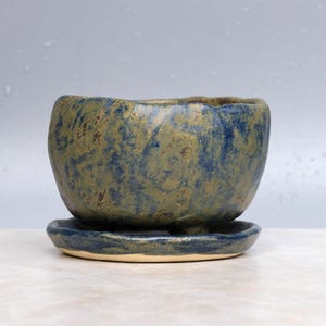 May include: A small, round, handmade ceramic planter with a textured surface featuring a mottled blend of deep blue and earthy olive green glaze. The pot sits securely on a matching circular ceramic saucer that shares the same blue and green finish.