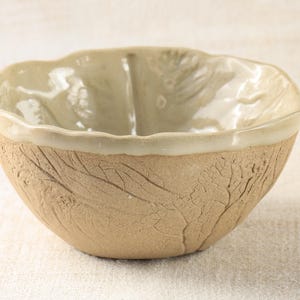 Puede incluir: Un pequeño cuenco de cerámica beige con un glaseado blanco en el interior y un exterior texturizado y rugoso. El cuenco tiene un borde ligeramente ondulado.