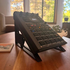 May include: A black Roland SP-404MKII sampler and effects processor mounted on a black angled stand. The device features a central circular display showing the text ON, numerous square rubber pads, and various control knobs and buttons.