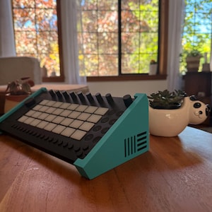 May include: A black and teal electronic music production device with a grid of white buttons and black knobs. The device is on a wooden surface, next to a white panda-shaped planter with a succulent plant.