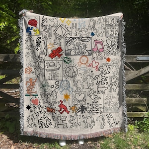 Op de afbeelding: Crèmekleurige sprei met een zwart en kleurrijk doodle-art ontwerp. De sprei toont verschillende figuren, symbolen en vormen, waaronder harten, muzieknoten en buitenaardse thema's. De randen hebben een franjes.