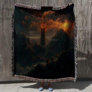 Puede incluir: Manta tejida que presenta un paisaje dram&aacute;tico con una alta torre bajo un cielo de fuego. El dise&ntilde;o incluye figuras silueteadas en una monta&ntilde;a, con una paleta de colores oscuros y bordes con flecos. La manta se muestra sobre un fondo neutro.