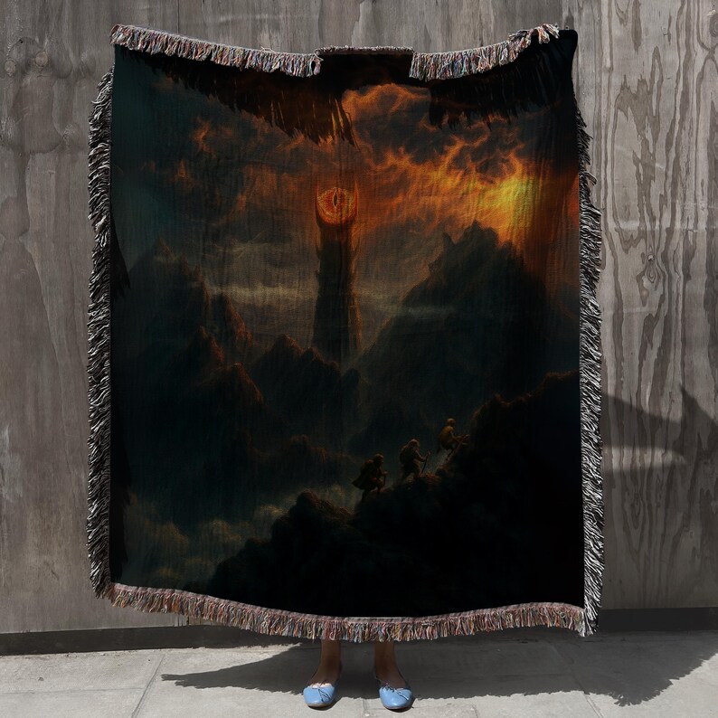 Puede incluir: Manta tejida que presenta un paisaje dram&aacute;tico con una alta torre bajo un cielo de fuego. El dise&ntilde;o incluye figuras silueteadas en una monta&ntilde;a, con una paleta de colores oscuros y bordes con flecos. La manta se muestra sobre un fondo neutro.